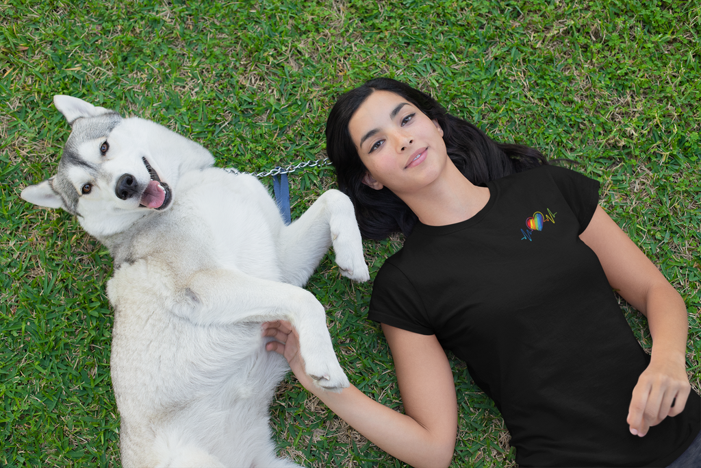 A women lays on the grass with her white dog. She has black hair and is staring at the camara with a small smile. She's wearing a blacks shirt with a rainbow heart and heart beat line embroidered. 