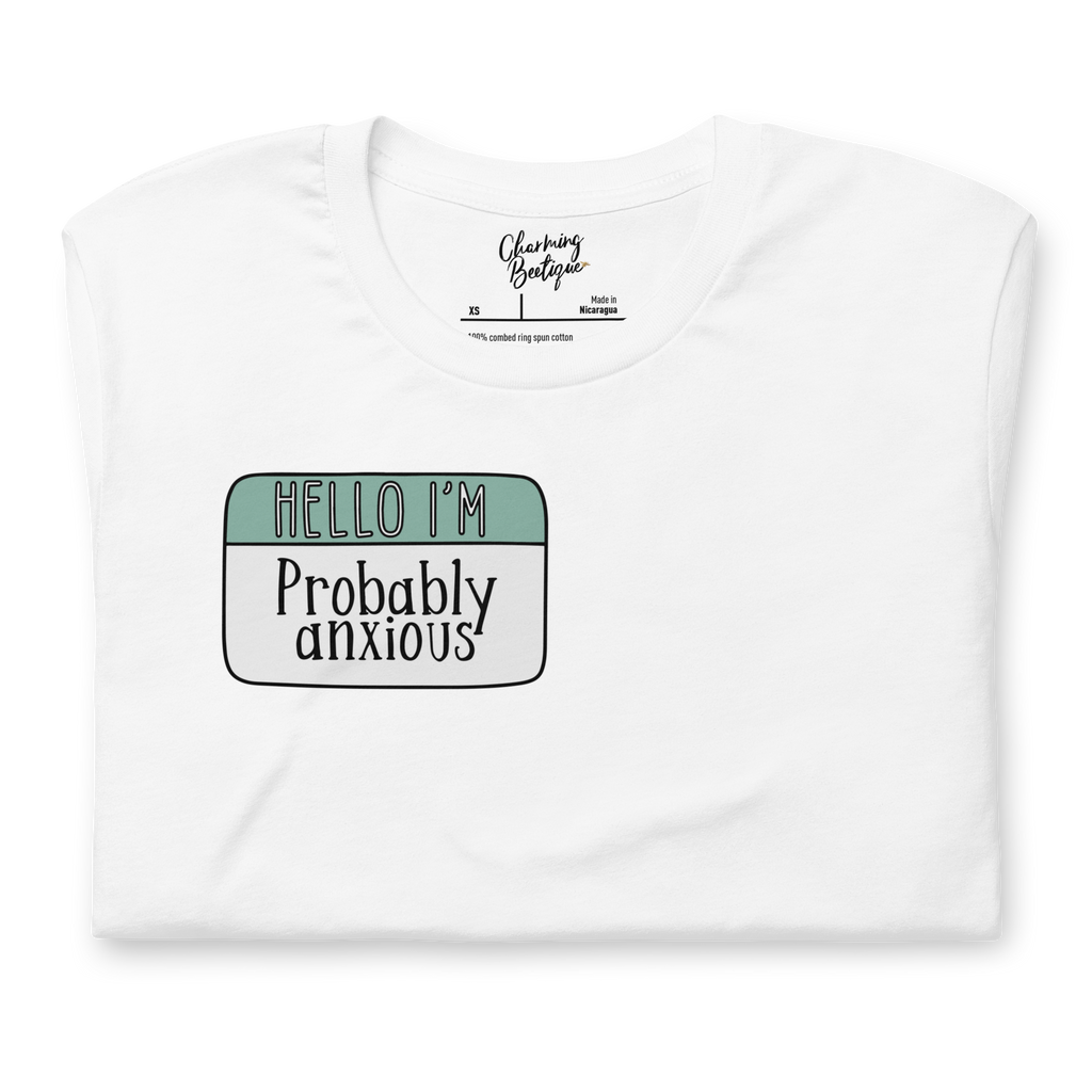 Image shows a white folded tshirt with a name tag graphic. The top of the name tag has text that reads 'hello I'm' light green; on the bottom of the name tag is off white with the word 'anxious'. 